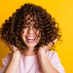 Mujer sonríe con pelo rizado y castaño, ideal para aprender como hidratar el pelo rizado y lucir así de radiante.