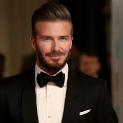 Hombre con pelo liso peinado hacia atrás, barba cuidada y esmoquin negro.