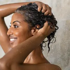 Mujer sonriente se lava el cabello rizado en la ducha, aplicando el método wavy para definir sus ondas.