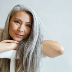 Mujer con cabello largo y canas amarillas, apoyada en una silla blanca.