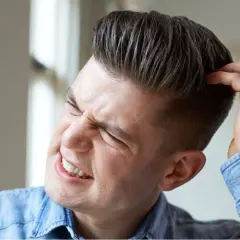 Hombre joven con expresión de molestia se rasca la cabeza, quizás por verrugas en la cabeza.