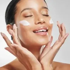 Mujer aplicando crema en su rostro, con los ojos cerrados y una sonrisa. La pregunta es: ¿que va primero la crema hidratante o el protector solar?