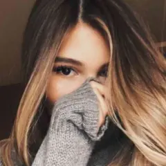 Chica con cabello largo y ondulado con mechas californianas, cubriendo parte de su rostro con un suéter gris.