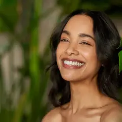 Sonrisa radiante de mujer con piel luminosa, rodeada de hojas verdes. El pantenol que es, un ingrediente clave para esta piel saludable.