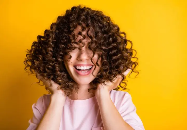 Mujer sonríe con pelo rizado y castaño, ideal para aprender como hidratar el pelo rizado y lucir así de radiante.
