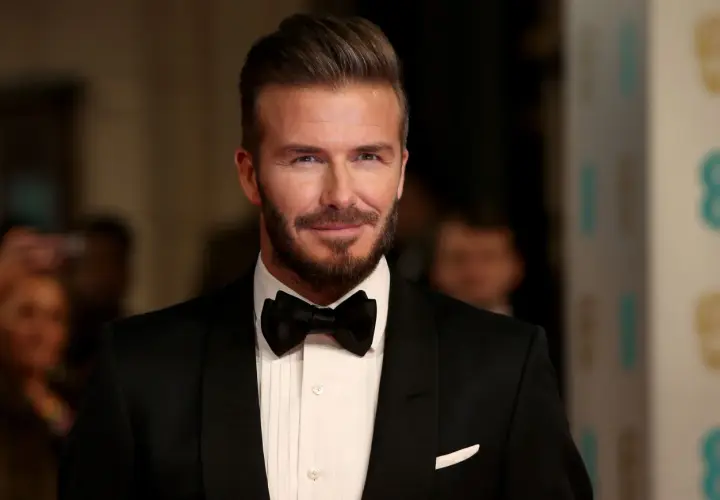 Hombre con pelo liso peinado hacia atrás, barba cuidada y esmoquin negro.
