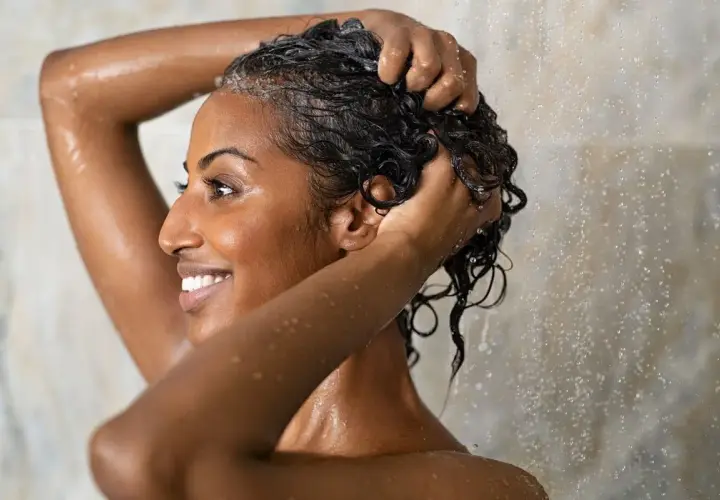 Mujer sonriente se lava el cabello rizado en la ducha, aplicando el método wavy para definir sus ondas.