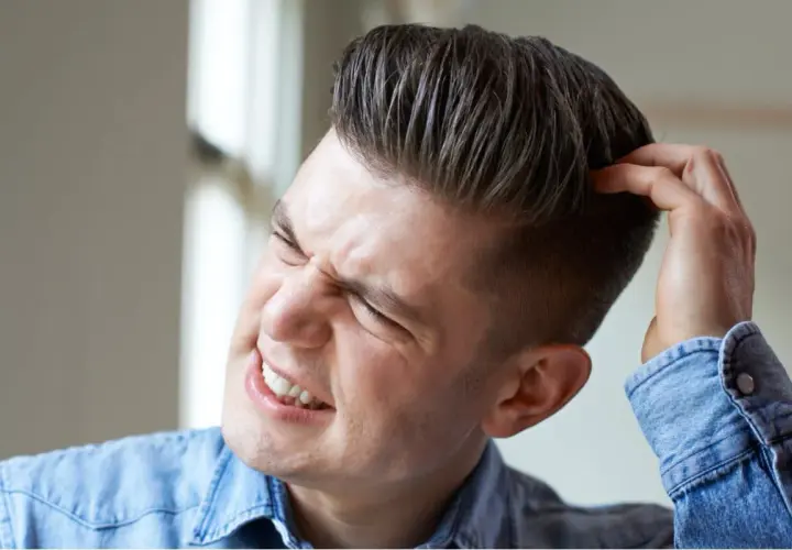 Hombre joven con expresión de molestia se rasca la cabeza, quizás por verrugas en la cabeza.