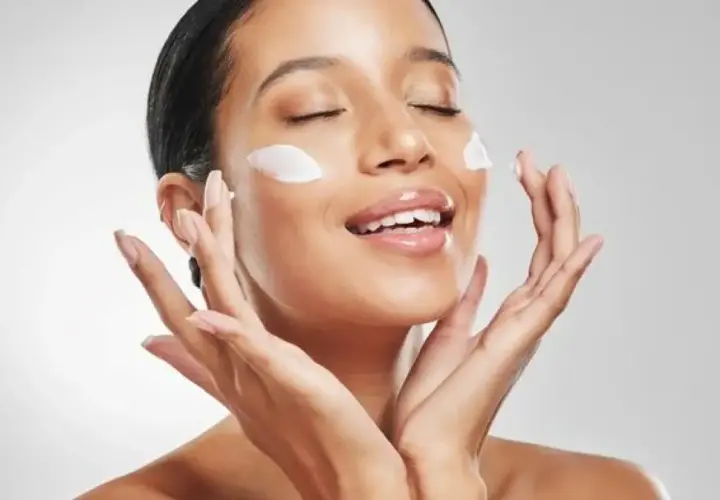 Mujer aplicando crema en su rostro, con los ojos cerrados y una sonrisa. La pregunta es: ¿que va primero la crema hidratante o el protector solar?