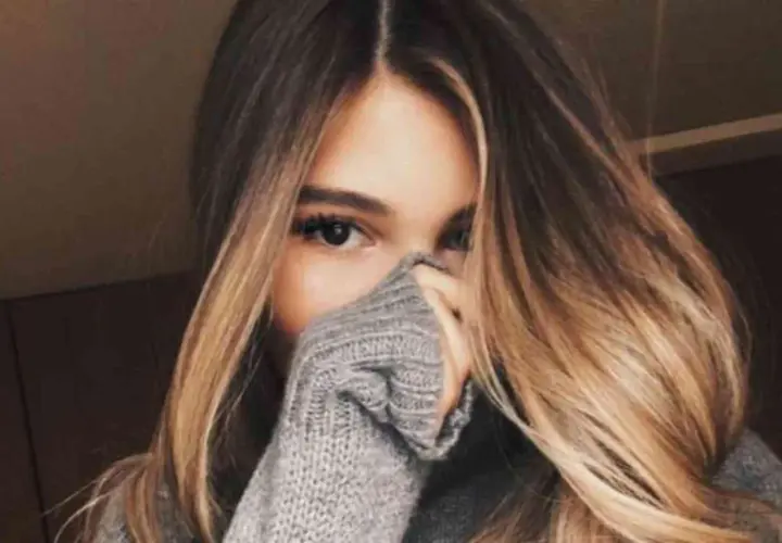 Chica con cabello largo y ondulado con mechas californianas, cubriendo parte de su rostro con un suéter gris.