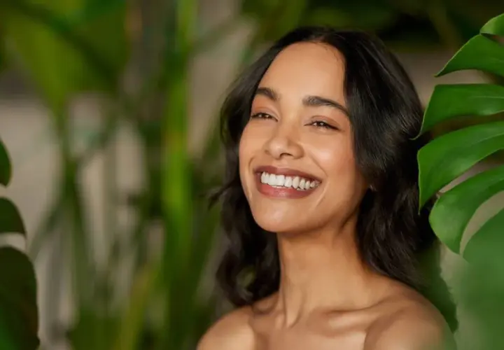 Sonrisa radiante de mujer con piel luminosa, rodeada de hojas verdes. El pantenol que es, un ingrediente clave para esta piel saludable.
