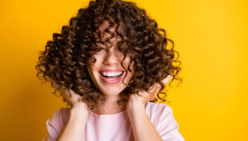 Mujer sonríe con pelo rizado y castaño, ideal para aprender como hidratar el pelo rizado y lucir así de radiante.