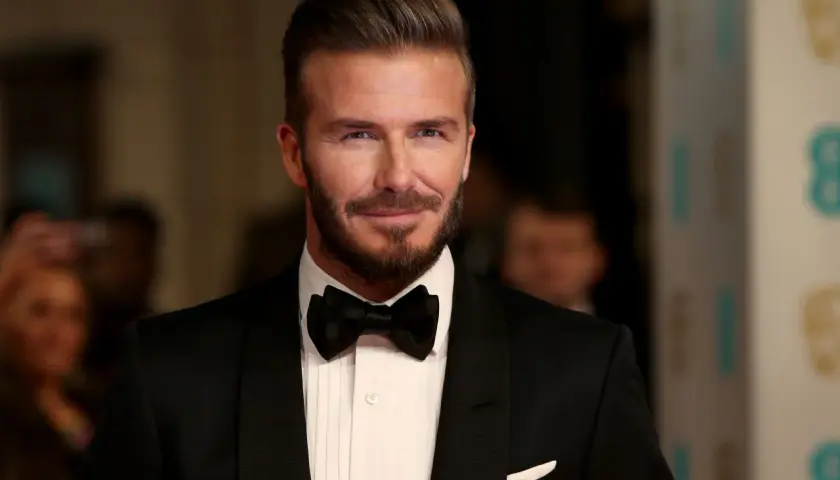 Hombre con pelo liso peinado hacia atrás, barba cuidada y esmoquin negro.