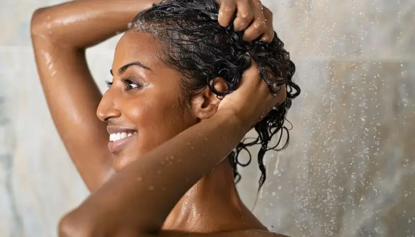 Mujer sonriente se lava el cabello rizado en la ducha, aplicando el método wavy para definir sus ondas.
