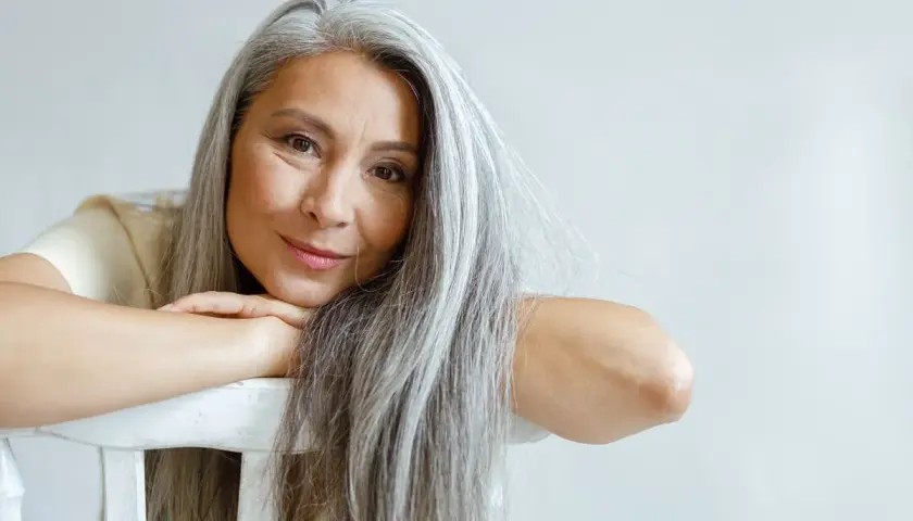Mujer con cabello largo y canas amarillas, apoyada en una silla blanca.