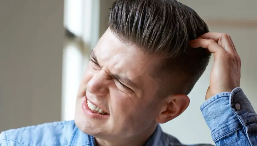Hombre joven con expresión de molestia se rasca la cabeza, quizás por verrugas en la cabeza.