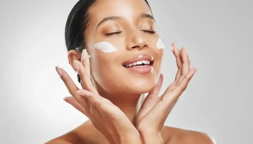 Mujer aplicando crema en su rostro, con los ojos cerrados y una sonrisa. La pregunta es: ¿que va primero la crema hidratante o el protector solar?
