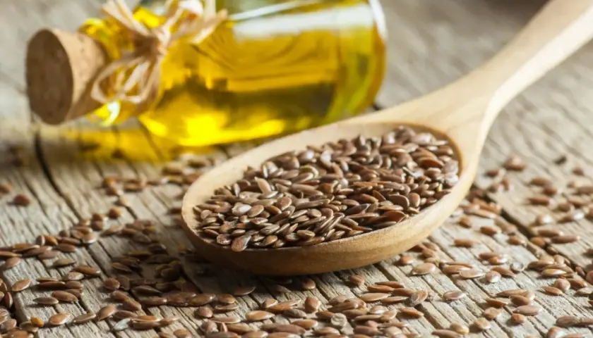 Cuchara de madera llena de semillas de lino, con una botella de aceite de lino al fondo. Descubre las increíbles aceite de lino propiedades.