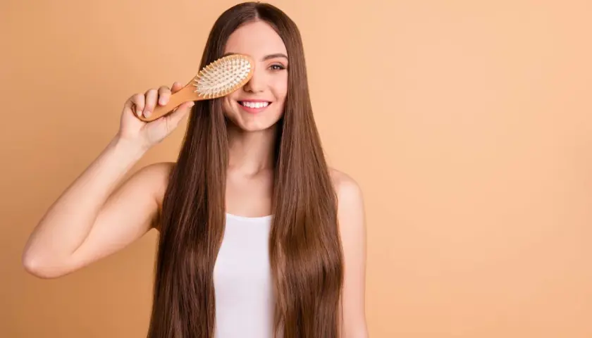 Mujer sonriente con pelo largo y liso, usando un cepillo de madera para mostrar cómo desenredar un pelo muy enredado.