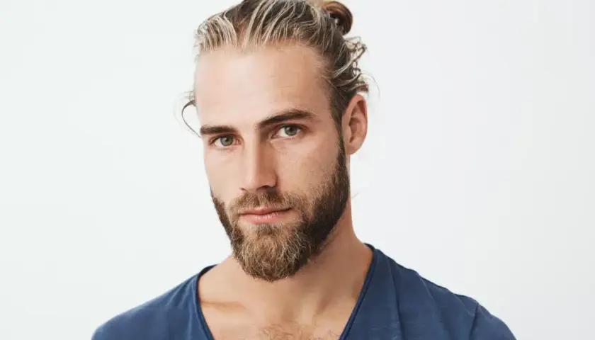Chico con barba y mechas rubias recogidas en un moño. Su mirada es intensa y directa.