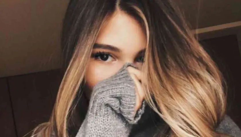Chica con cabello largo y ondulado con mechas californianas, cubriendo parte de su rostro con un suéter gris.