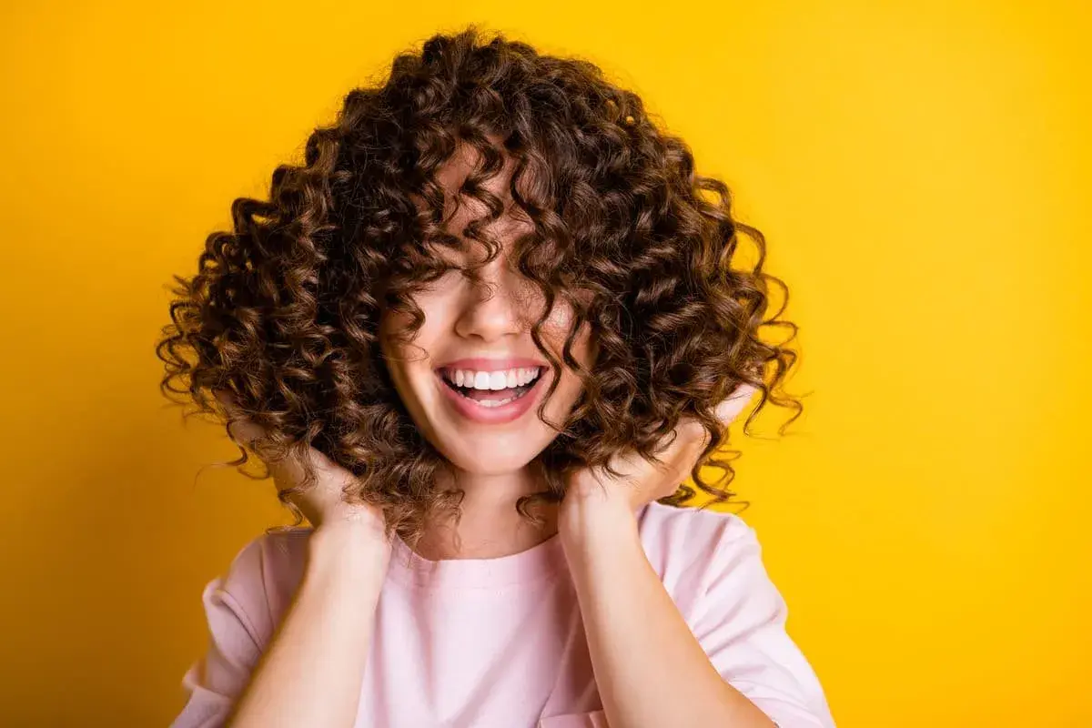 Mujer sonríe con pelo rizado y castaño, ideal para aprender como hidratar el pelo rizado y lucir así de radiante.