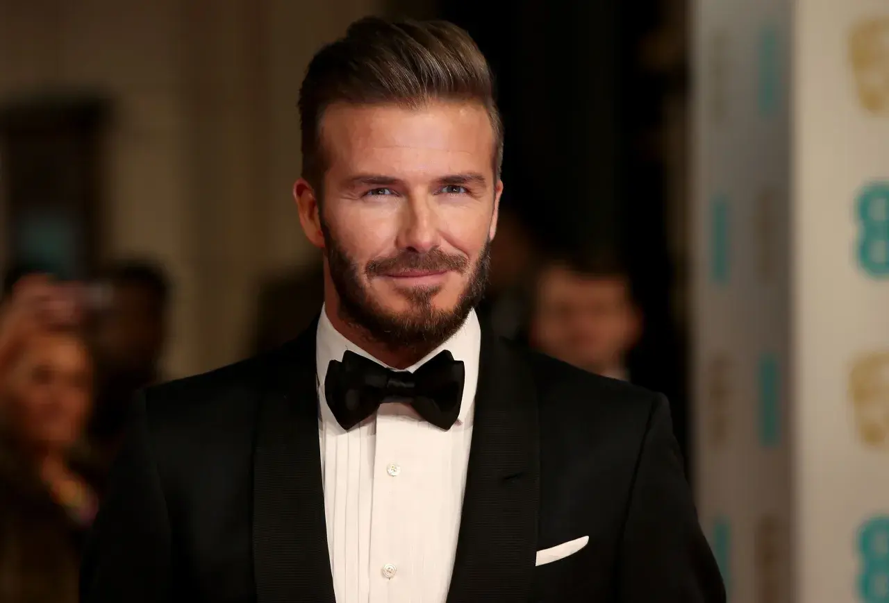 Hombre con pelo liso peinado hacia atrás, barba cuidada y esmoquin negro.