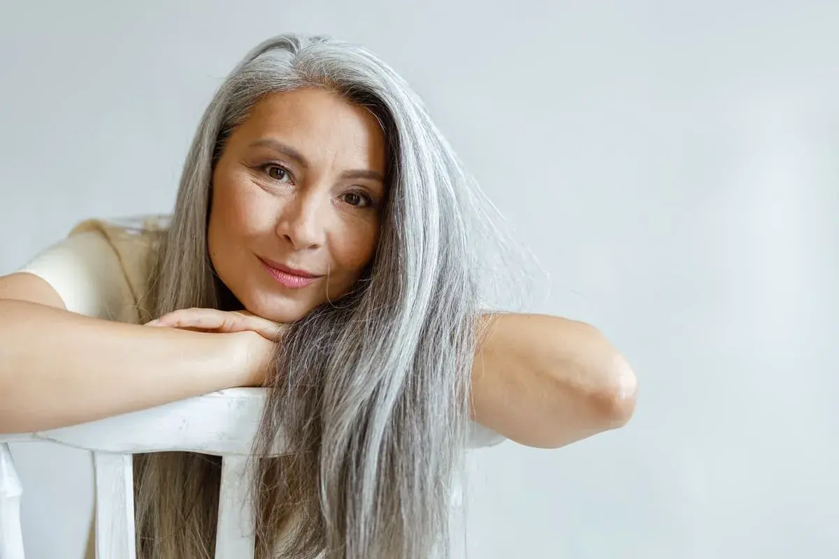 Mujer con cabello largo y canas amarillas, apoyada en una silla blanca.