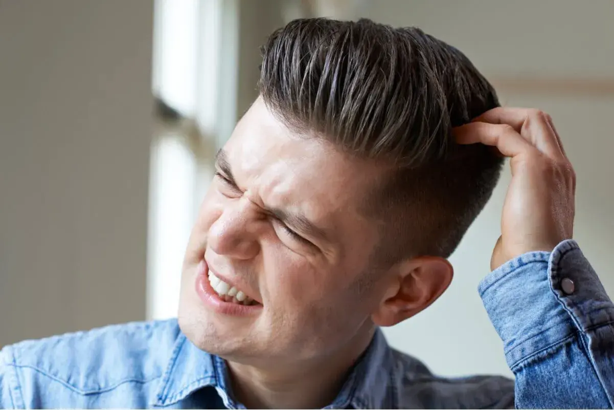 Hombre joven con expresión de molestia se rasca la cabeza, quizás por verrugas en la cabeza.