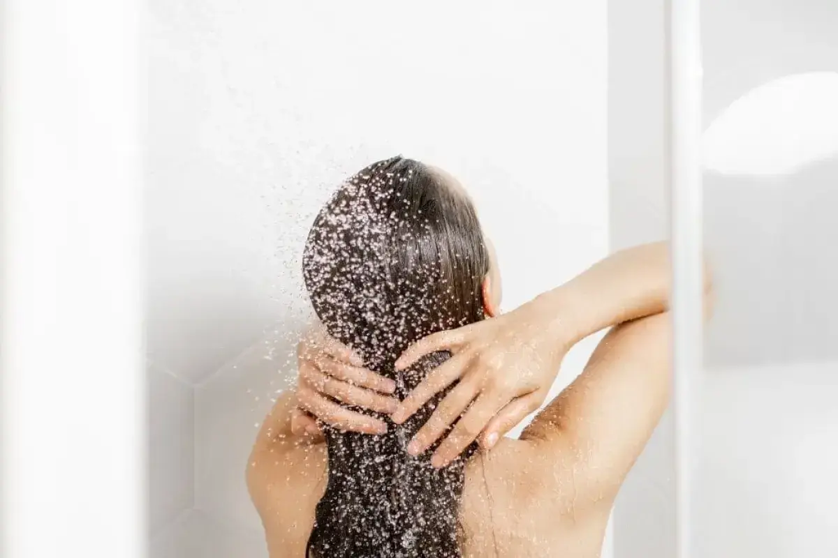 Persona disfrutando de un refrescante lavado de pelo bajo la ducha, con el agua cayendo sobre su cabello oscuro y las manos masajeando suavemente.