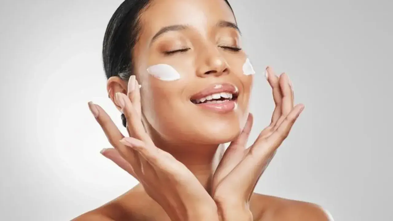 Mujer aplicando crema en su rostro, con los ojos cerrados y una sonrisa. La pregunta es: ¿que va primero la crema hidratante o el protector solar?