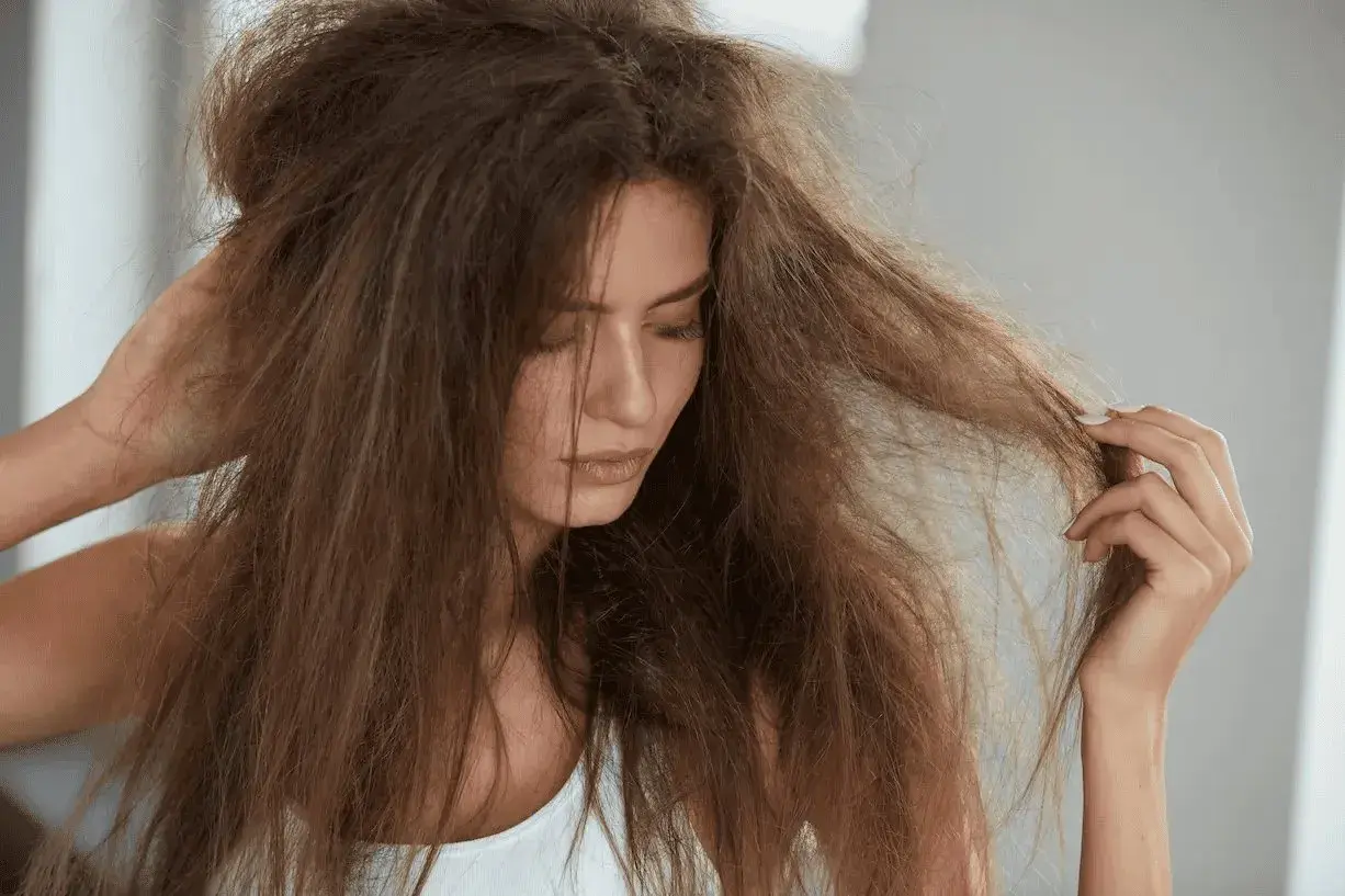 Mujer con pelo fosco y enredado, tocándose la cabeza con una mano y examinando un mechón con la otra.