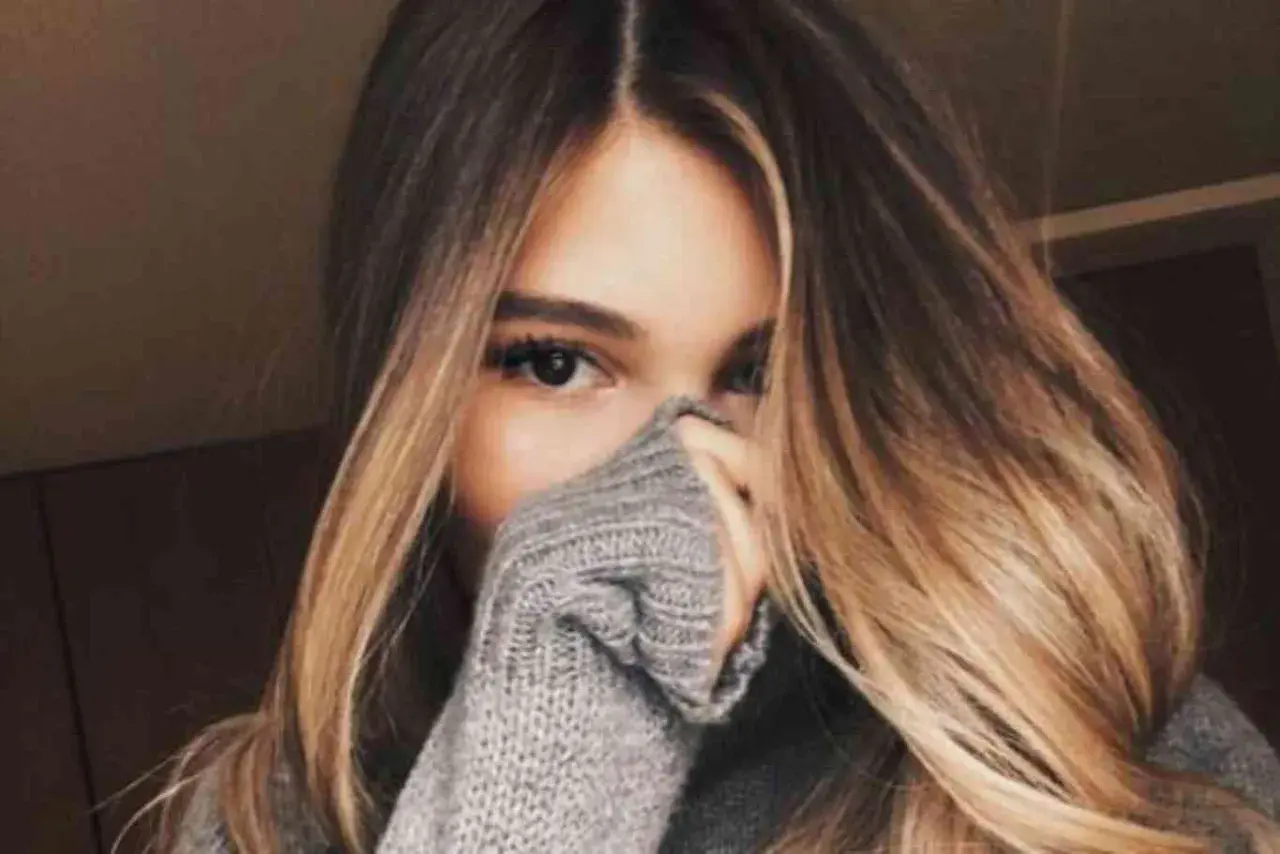 Chica con cabello largo y ondulado con mechas californianas, cubriendo parte de su rostro con un suéter gris.