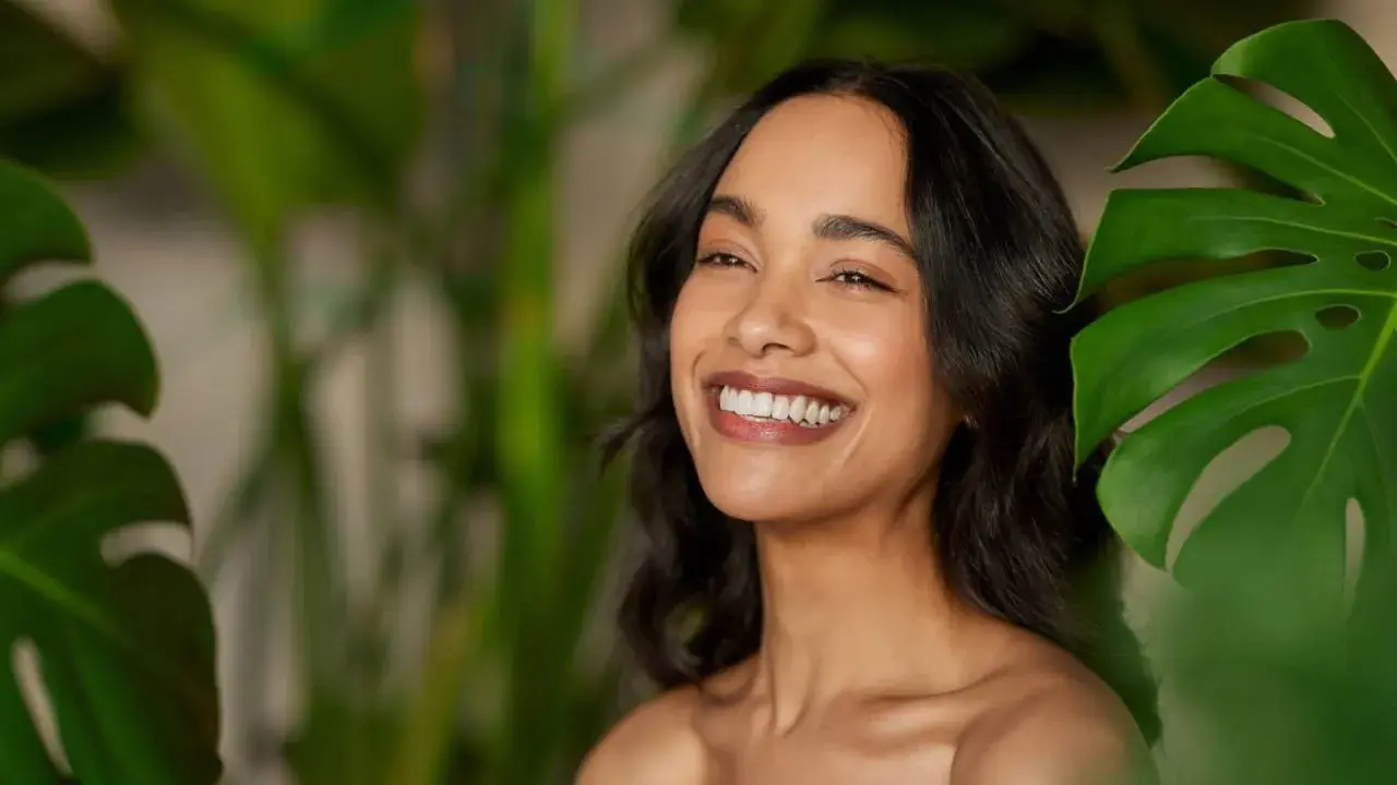 Sonrisa radiante de mujer con piel luminosa, rodeada de hojas verdes. El pantenol que es, un ingrediente clave para esta piel saludable.
