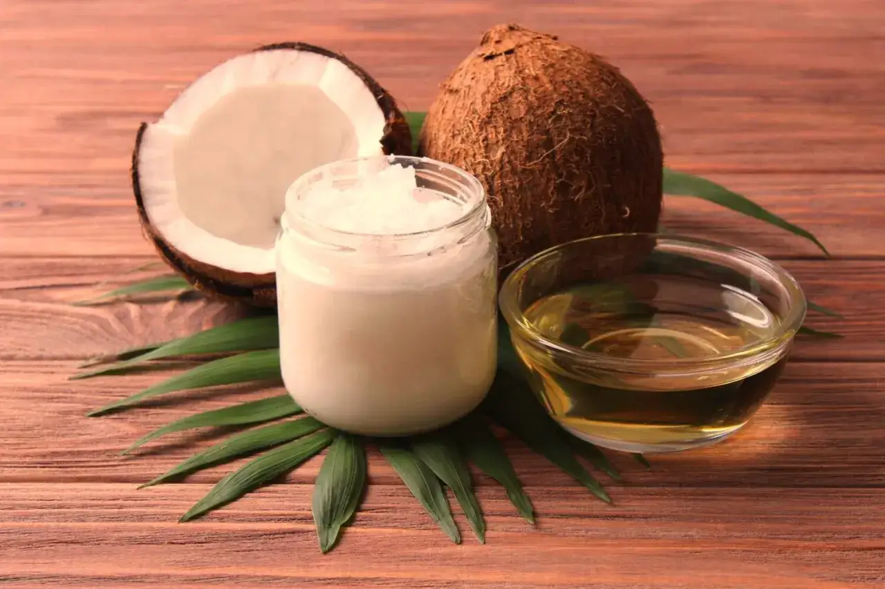 Aceite de coco para que sirve: un coco abierto, un frasco de aceite de coco sólido y un cuenco de aceite de coco líquido sobre hojas verdes.