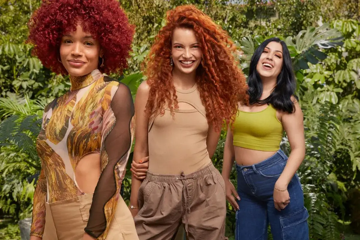 Tres mujeres posan sonrientes, una con rizos rojos vibrantes, otra con cabello pelirrojo ondulado y la tercera con cabello oscuro. Todas lucen atuendos modernos, destacando la diversidad de tonos de pelirrojo natural.