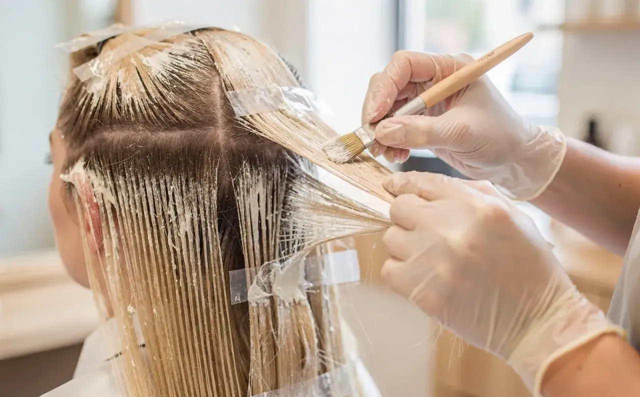 Manos enguantadas aplican tinte a las mechas rubias. Se pregunta cuánto duran las mechas.