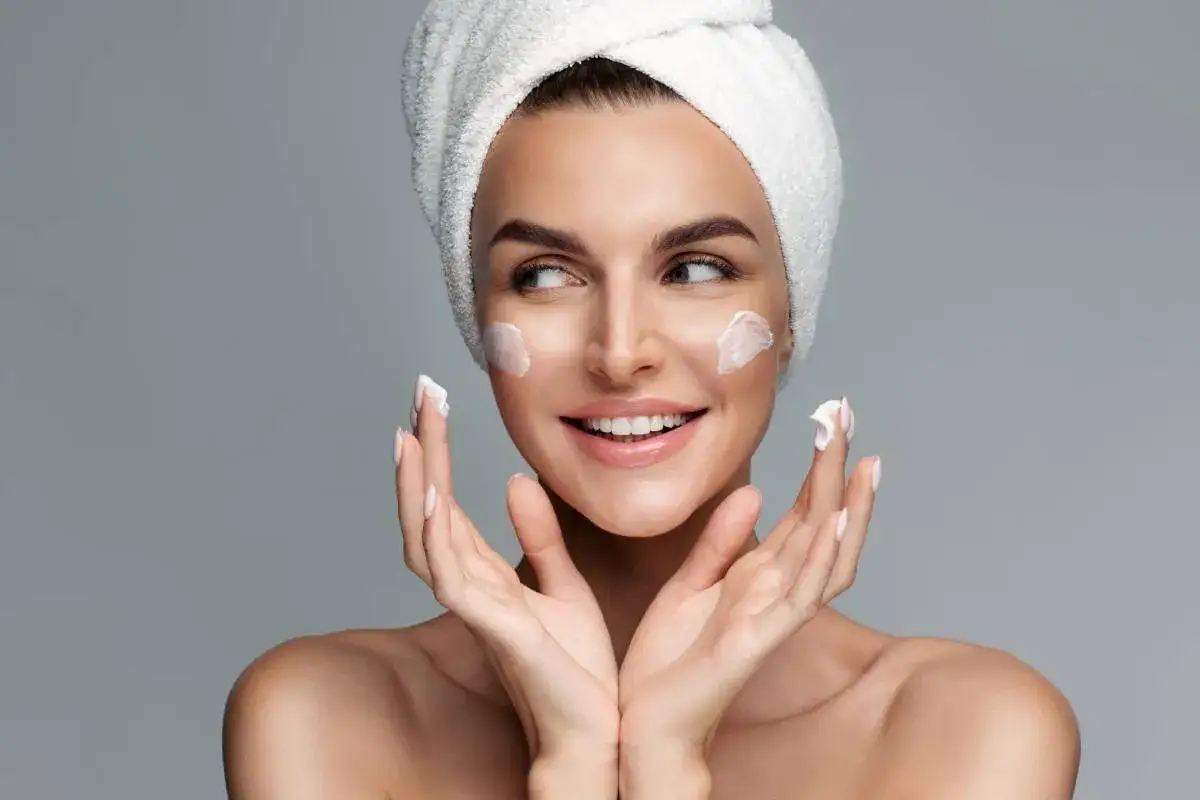 Mujer con toalla en la cabeza y crema en las mejillas, cuidando la elasticidad de la piel.