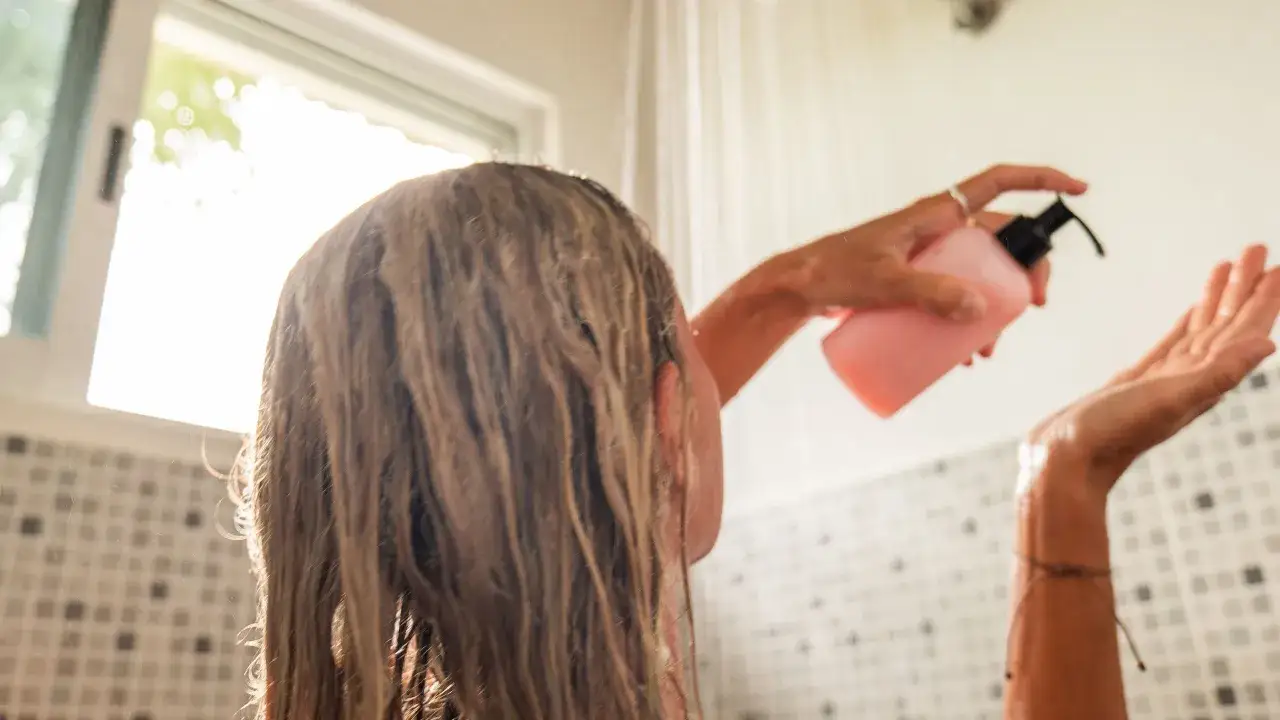 Persona aplica acondicionador en su cabello mojado bajo la ducha, preparándose para que el acondicionador se enjuaga.
